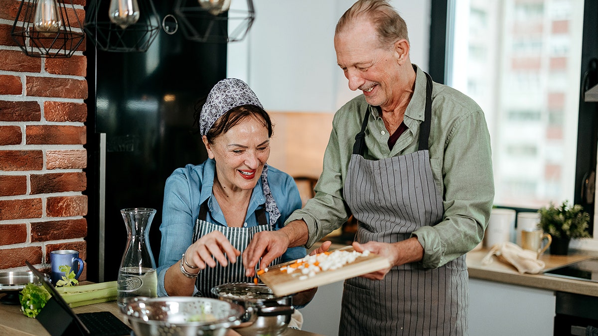An older couple cooking together in a modern kitchen, smiling while adding chopped vegetables to a pan.