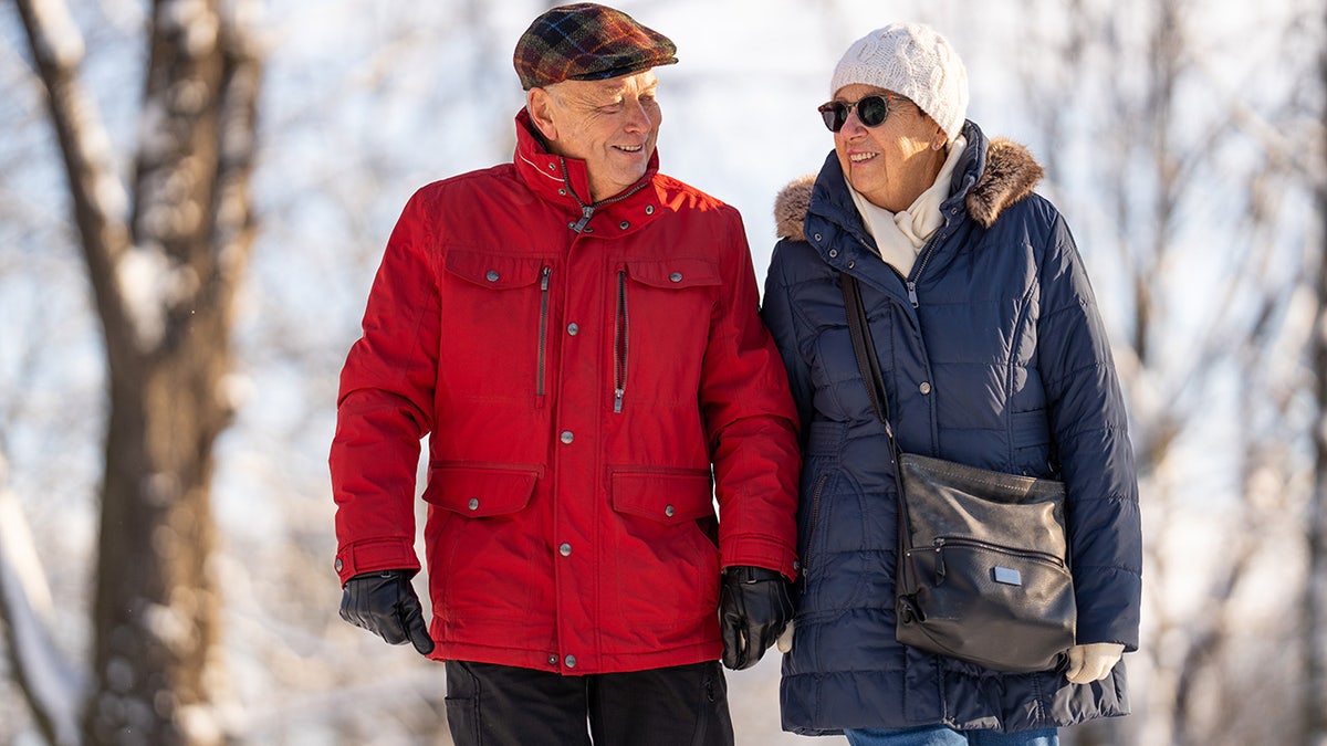 Senior couple bundled up in winter smiling and enjoying winter exercise walk with trees in background.