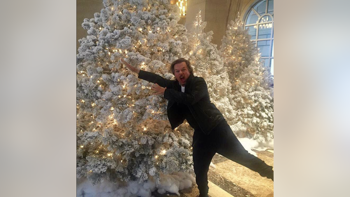 David Spade strikes a funny pose in front of a Christmas tree.