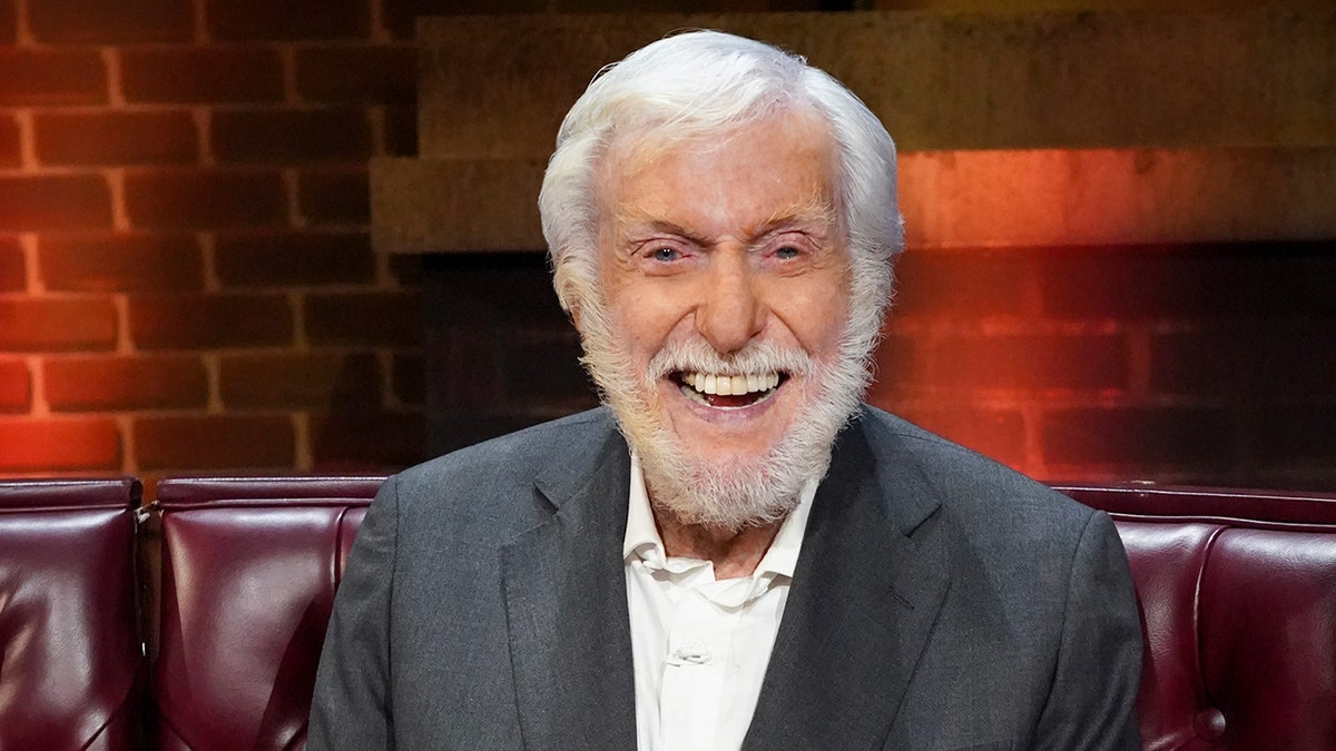 Dick Van Dyke sitting on a oxblood red couch and smiling wearing a grey suit.