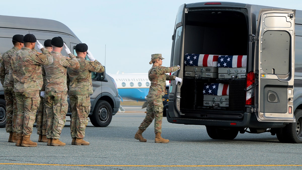 A soldier shuts the rear door of a transport vehicle holding multiple flag-draped transfer cases on an airfield.