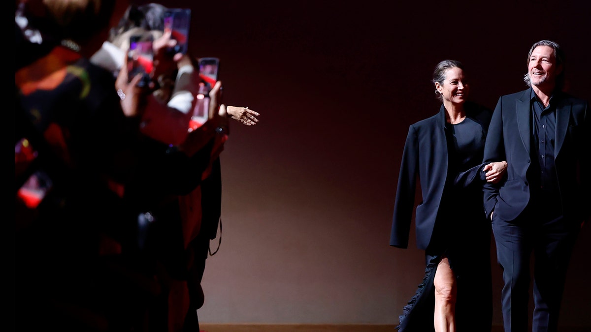 Ed Burns and Christy Turlington walking in front of cameras and smiling on the red carpet.