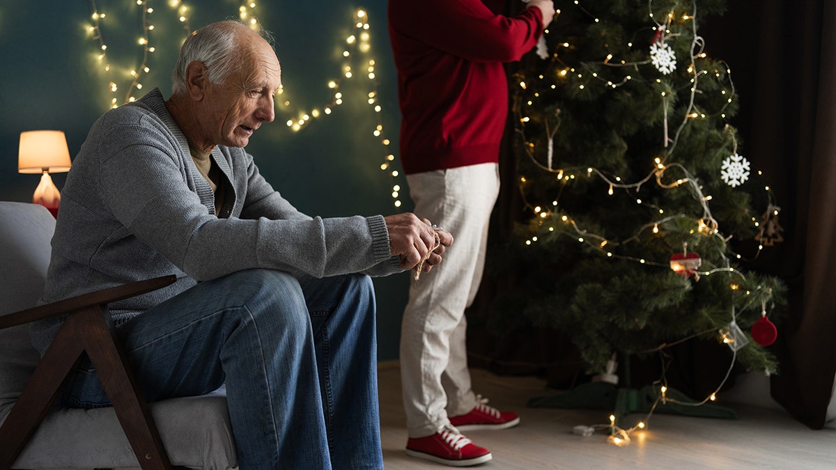 Elderly man sitting, looking sad and lonely as son in background decorates Christmas tree.