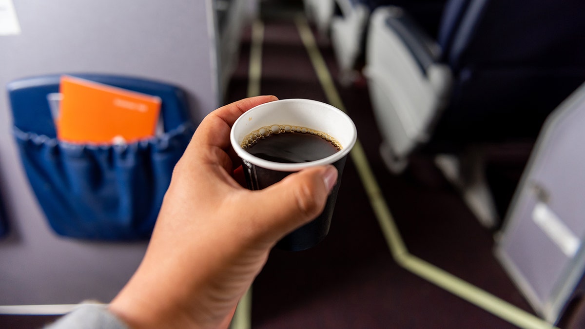 Hand holding a small cup of hot black coffee inside an airplane cabin with seat-back pockets and rows of seats visible.