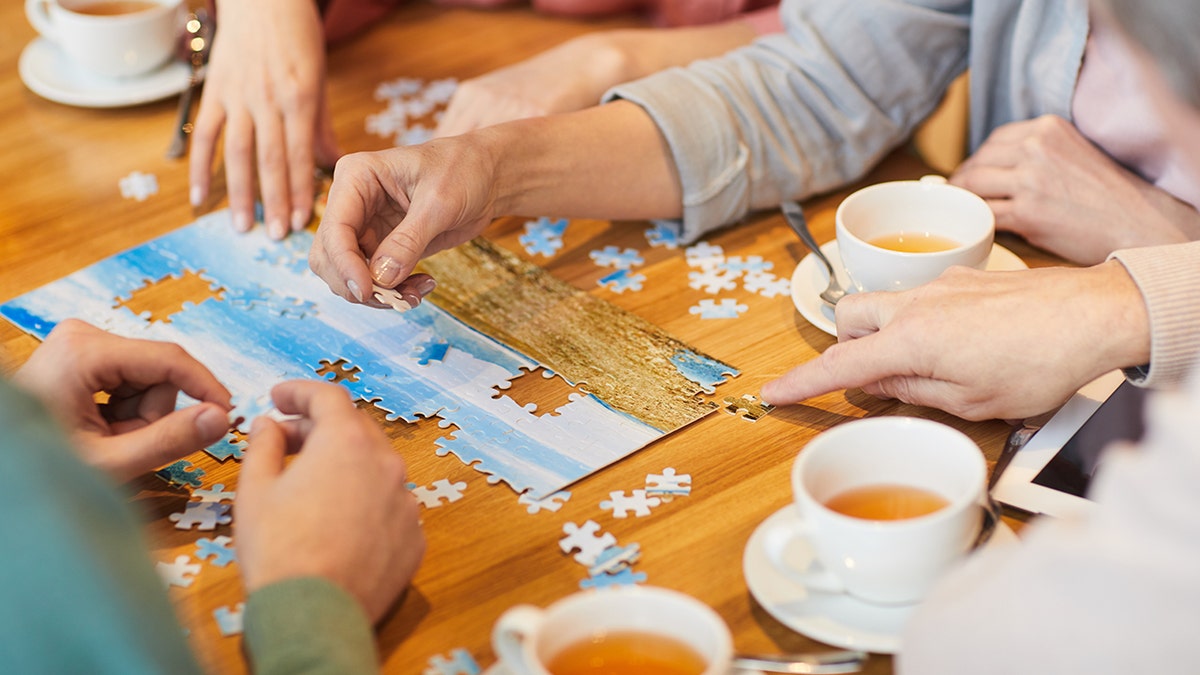 People working together on a jigsaw puzzle at a wooden table with cups of tea nearby.