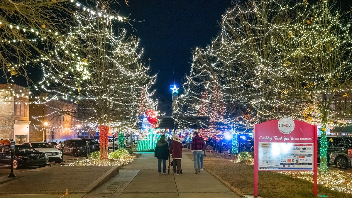 People walking through a downtown street lined with trees wrapped in white Christmas lights during a festive holiday celebration at night.