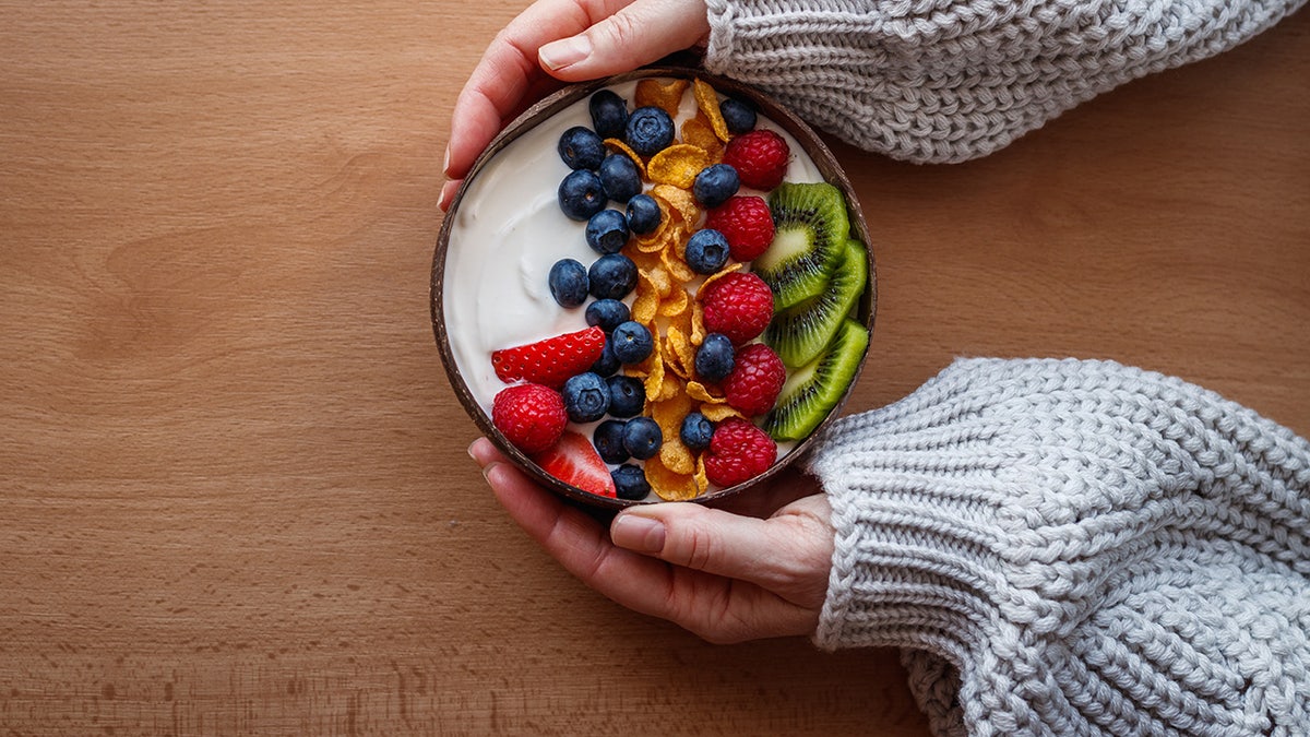 Woman wearing thick, cozy sweater holds bowl of yogurt tipped with fruit.