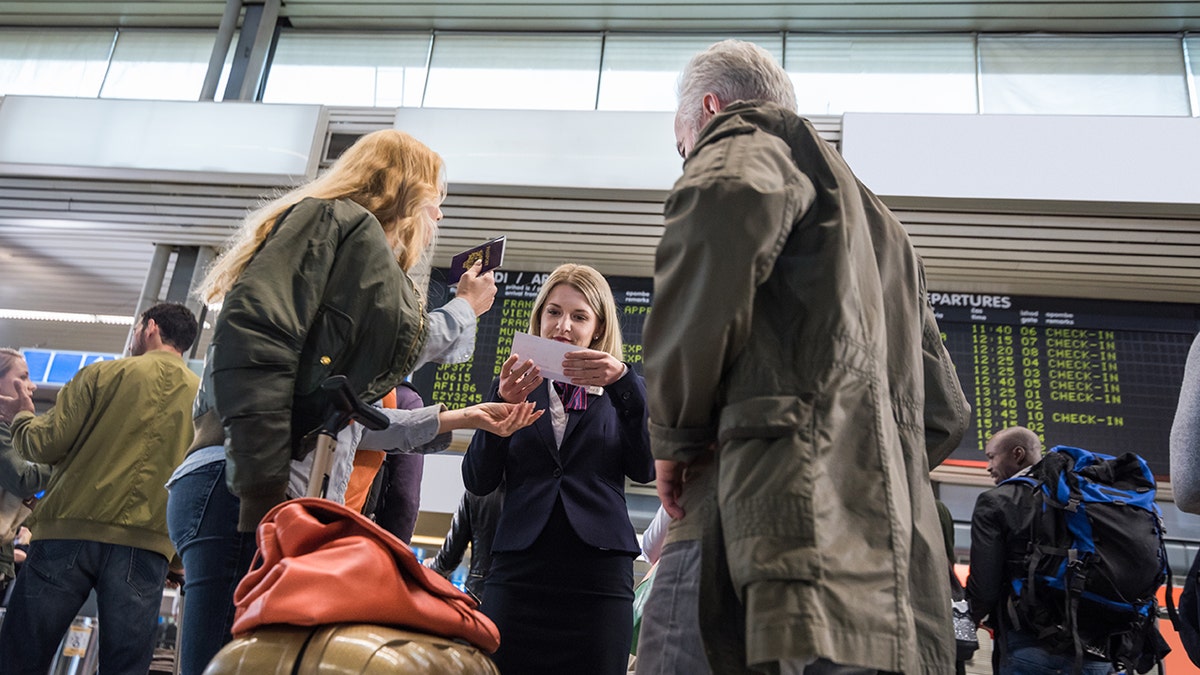 Airline staff assisting travelers at an airport check-in area as passengers present documents near a departures board.