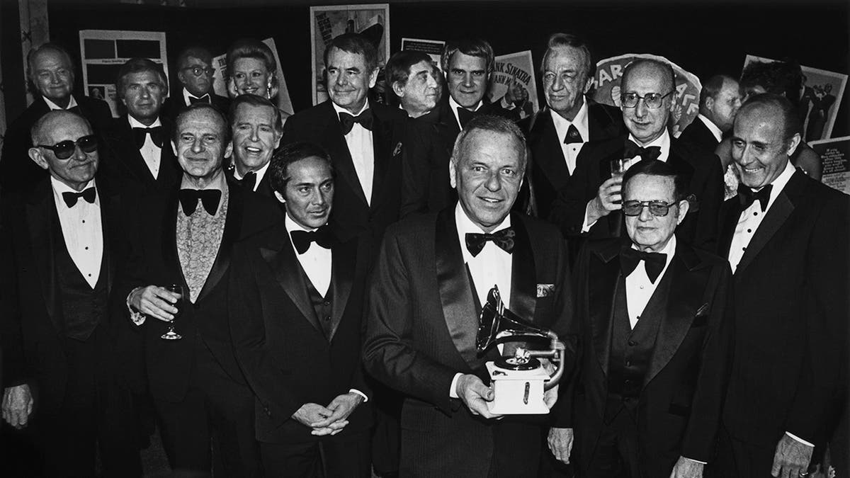 Frank Sinatra holding an award surrounded by men in suits as they all smile.