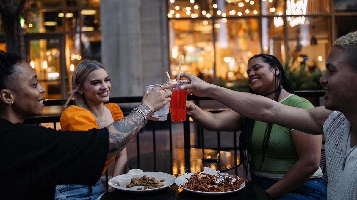Young, diverse group of friends dining in early evening outdoors, toasting drinks with food seen on table.
