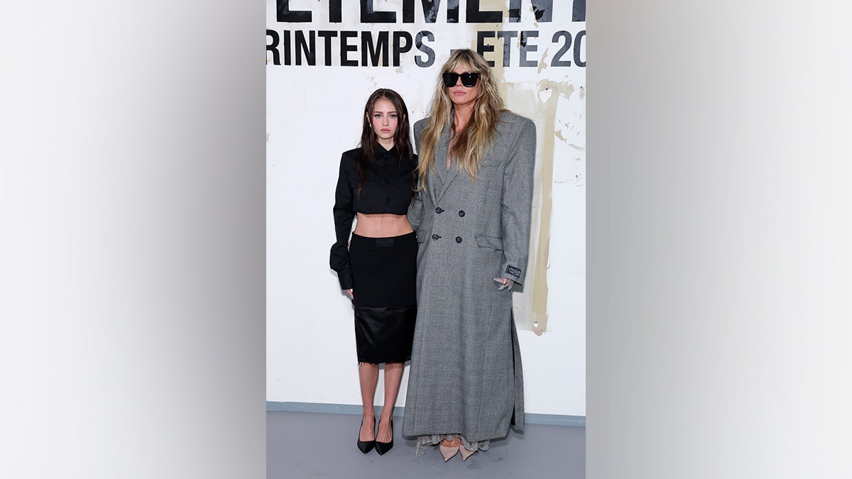 Heidi Klum and her daughter Leni Klum pose together at Paris Fashion Week, with Klum wearing a long gray tailored coat and Leni wearing a black cropped top and skirt.