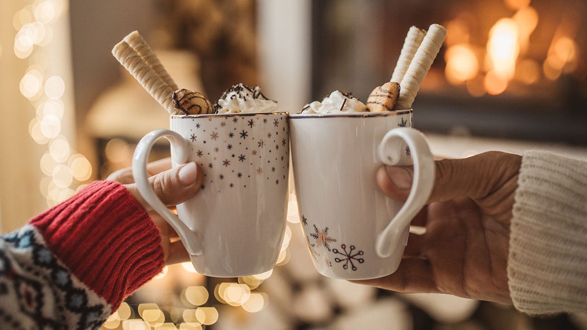 Man and woman's hands holding mugs of hot chocolate loaded with toppings in front of fireplace.