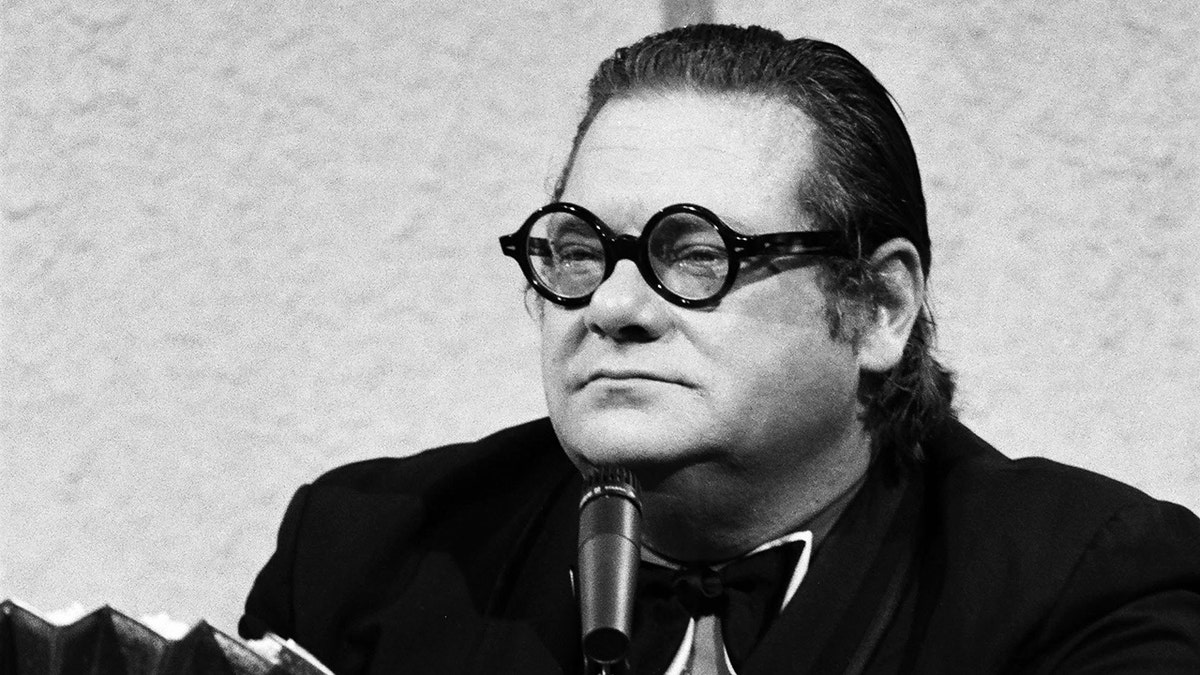 Gambar close-up Jackie Vernon mengenakan setelan gelap dengan kacamata bundar.