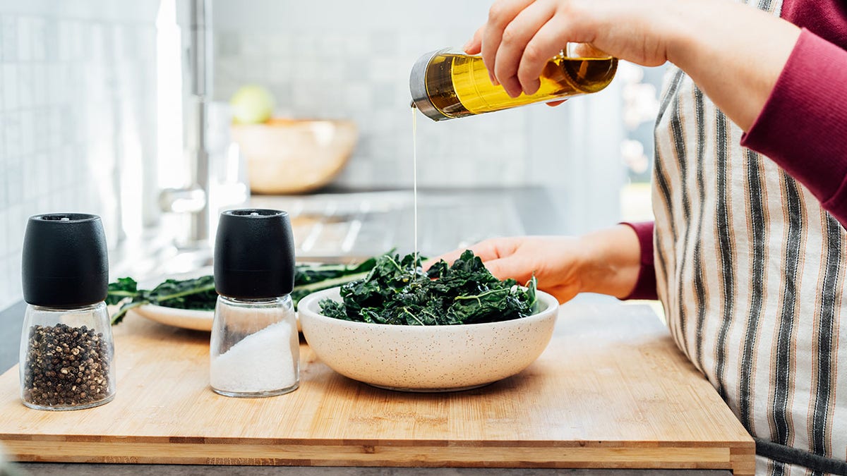 Womans hand seen pouring olive oil into salad bowl of kale with salt and pepper shakers on side.