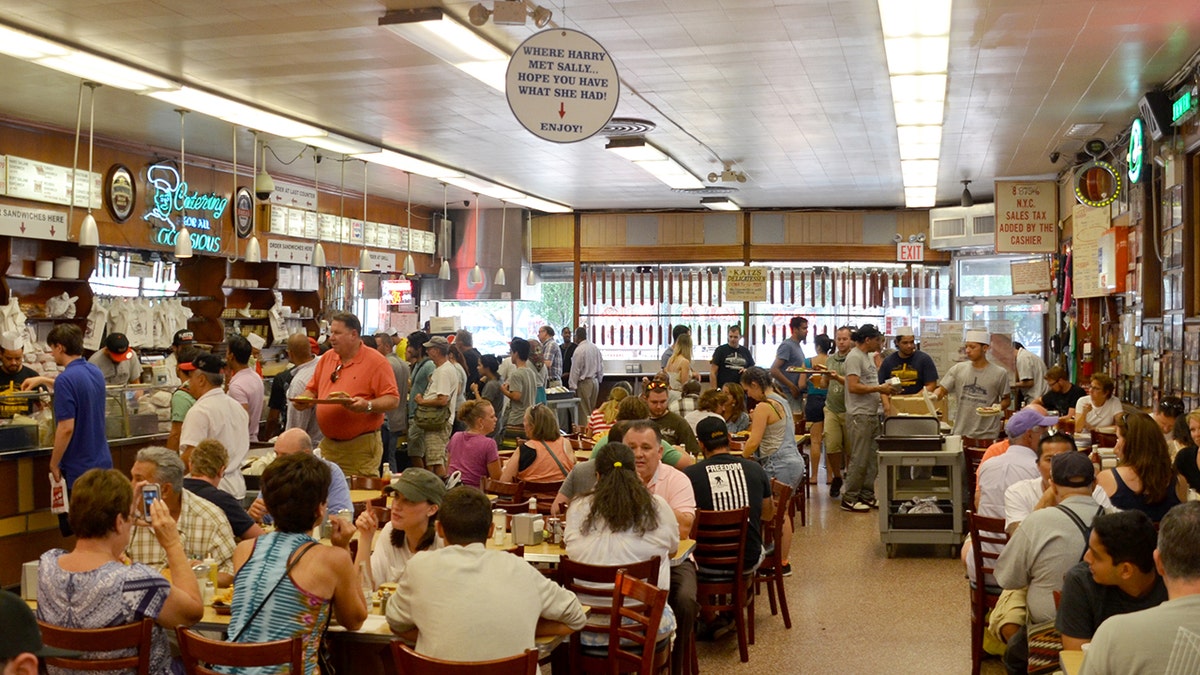 Busy packed Katz's Deli with sign seen hanging from ceiling that says "Where Harry met Sally" with red arrow pointing to table where iconic movie seen was filmed.