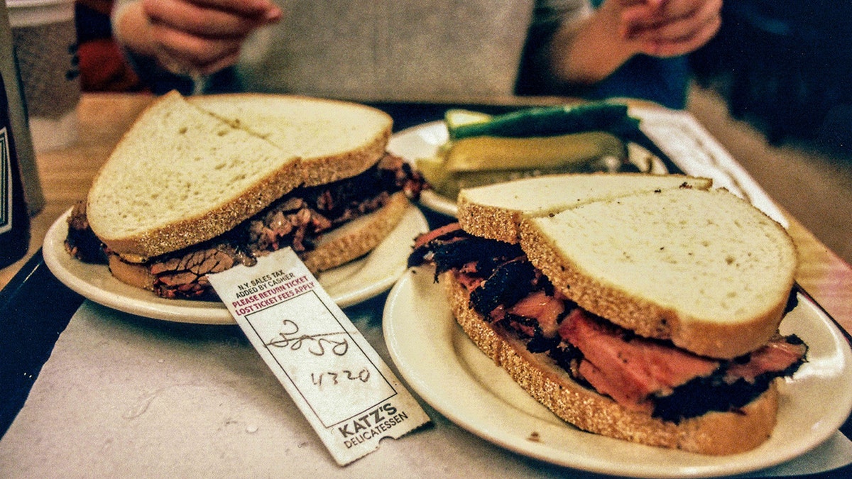 An order ticket lies amongst pastrami on rye and brisket on rye sandwiches with a plate of pickles at Katz's Delicatessen in Manhattan, New York, USA. Person's hands seen in background as they prepare to eat.