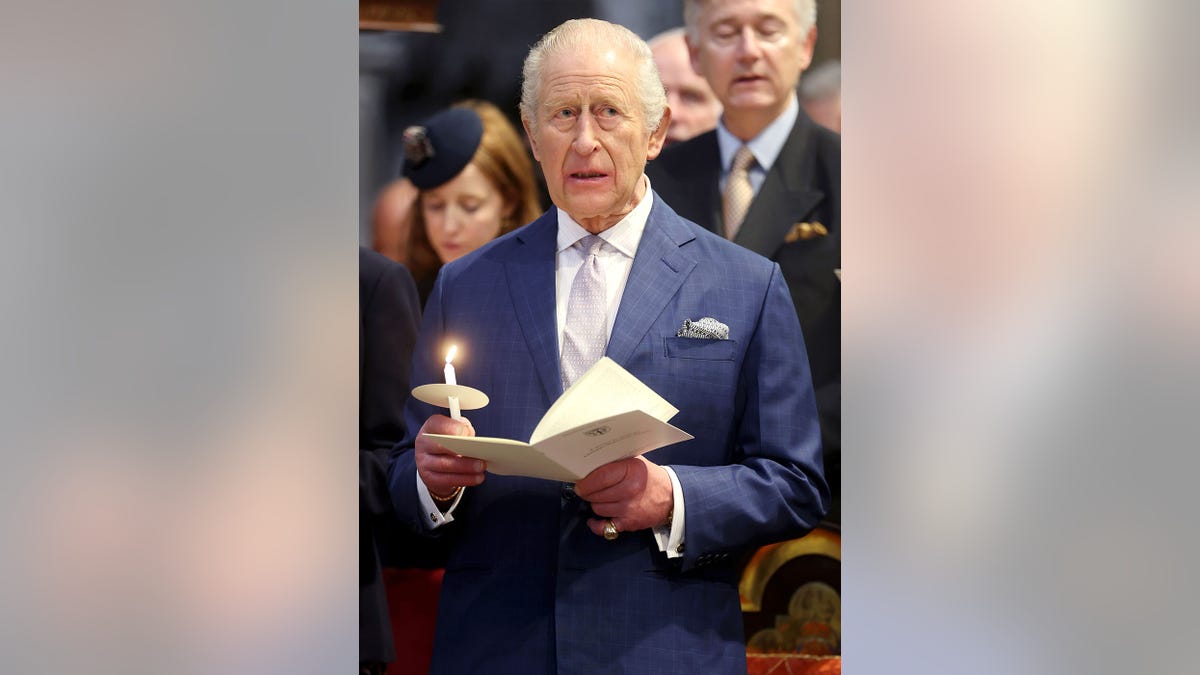 King Charles holding a candle and a prayer book while wearing a blue suit.