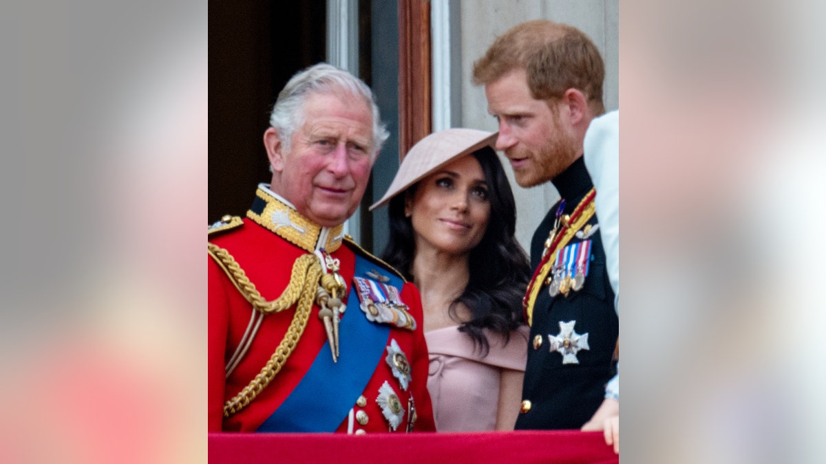 Meghan Markle looks at Prince Harry as King Charles, in a royal red military suit, looks on from the Buckingham Palace balcony.