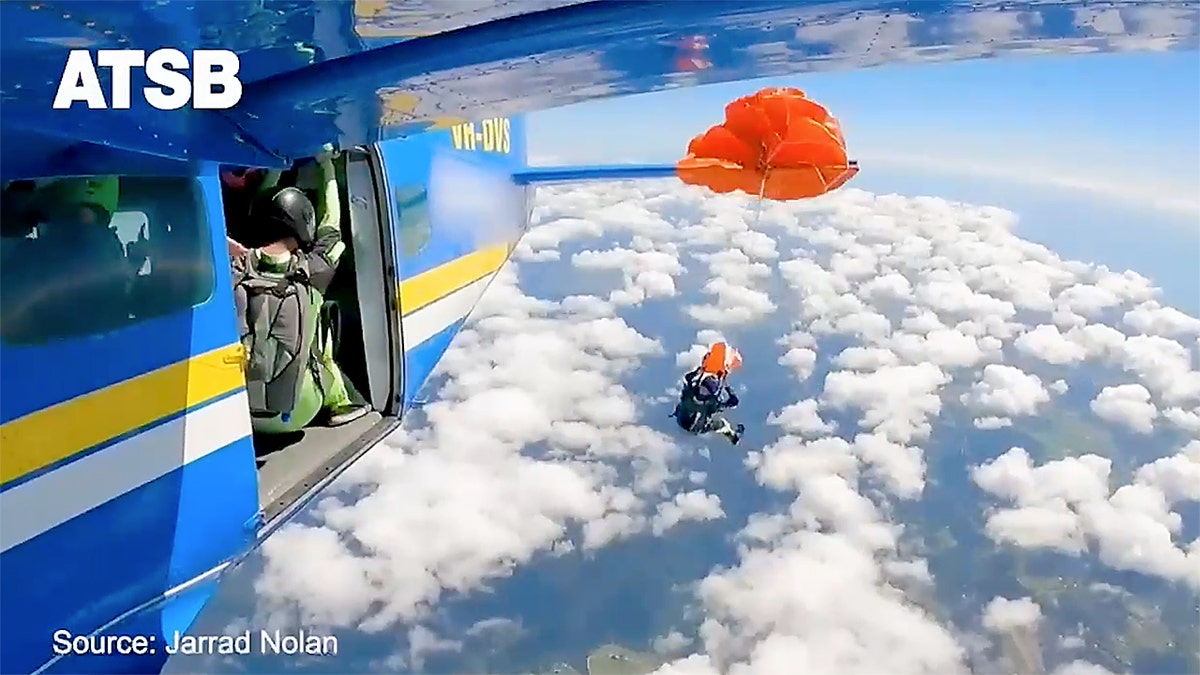 A skydiver drifts beneath a partially opened orangish reserve parachute that is caught connected the aircraft's wing.