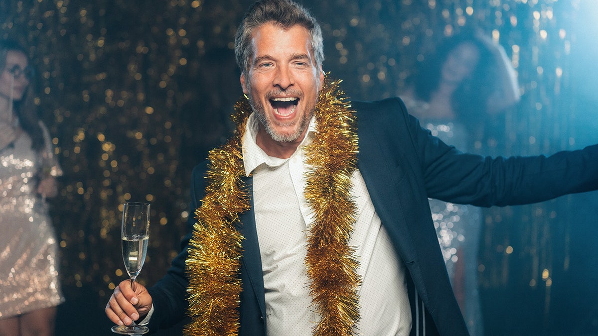 Man dancing, smiling on New Year's Eve, wearing suit and gold garland around neck, holding glass of sparkling wine.