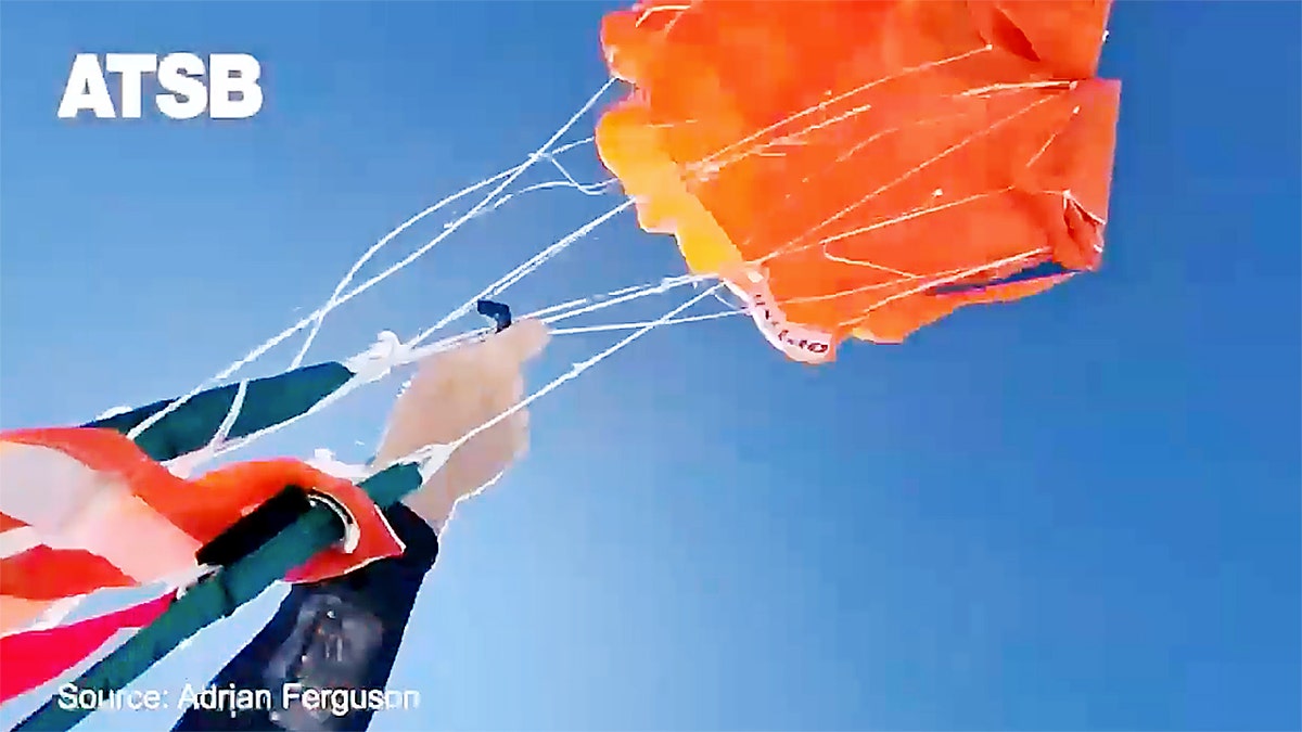 A skydiver grips parachute lines arsenic an orangish reserve canopy inflates against a wide bluish sky.