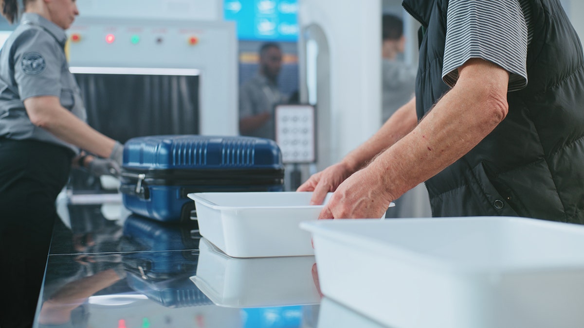 Airport security screening area with a passenger placing personal items in a tray while TSA agents inspect luggage at an X-ray checkpoint.