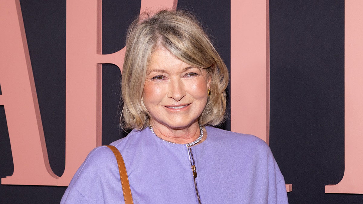 Older woman with shoulder-length blonde hair wearing a lavender top and gold earrings, smiling at a formal event with a dark backdrop.