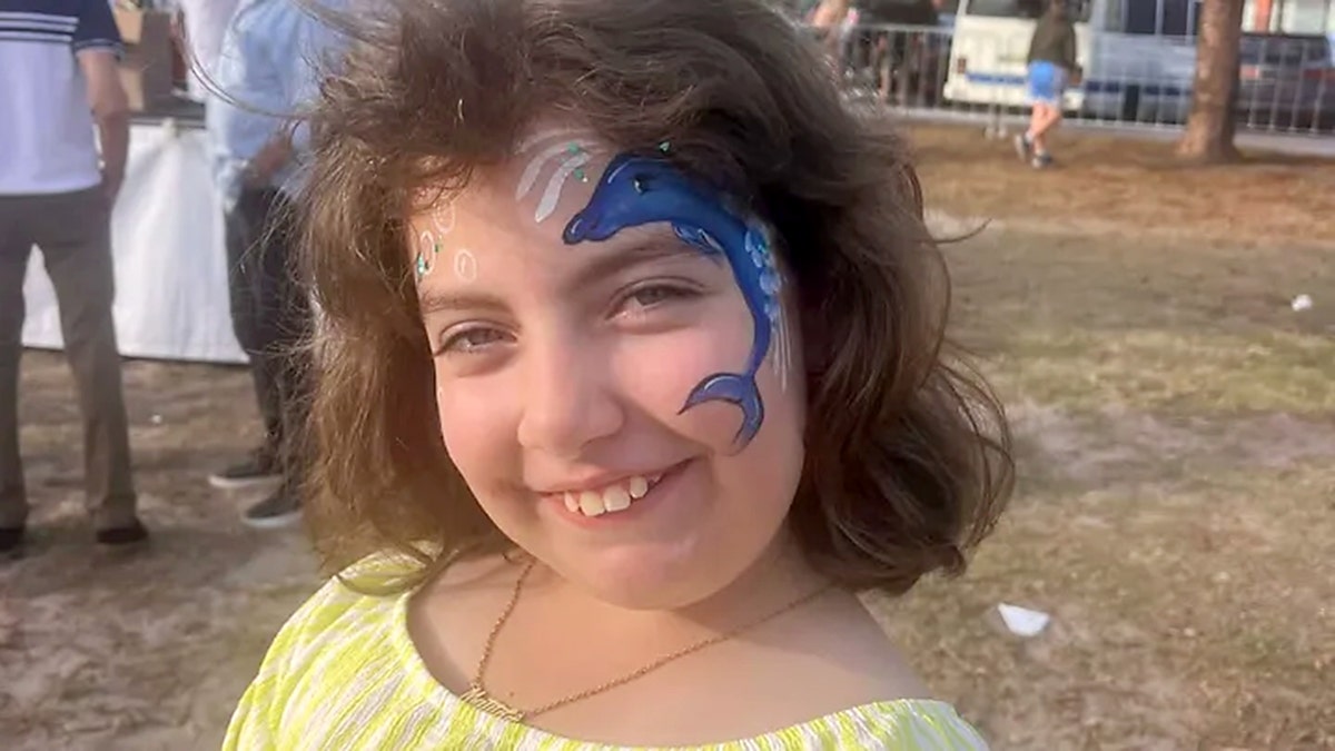 Smiling child with dolphin face paint stands outdoors at a daytime event.