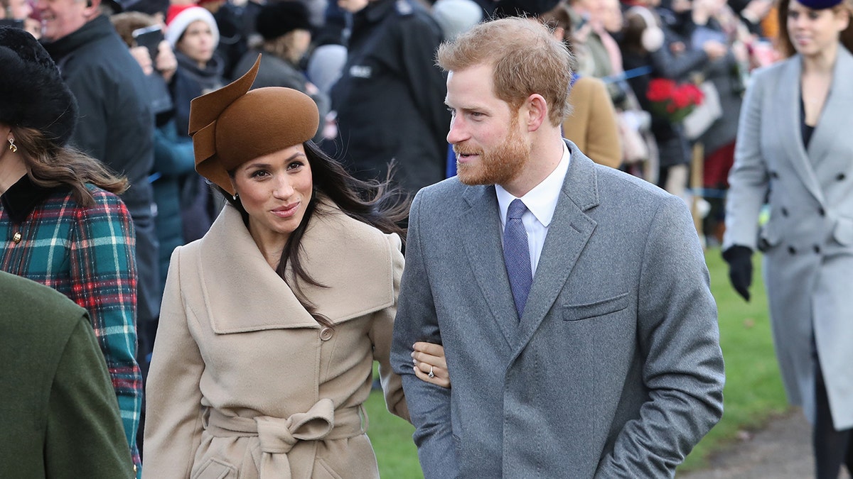 Meghan Markle wearing a brown hat and matching jacket on the arm of Prince Harry in a grey suit.