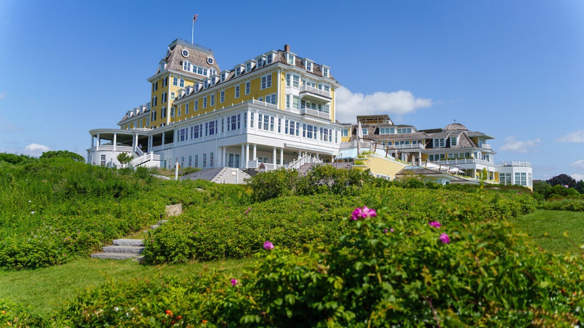 Ocean House hotel in Watch Hill, painted yellow and white, standing atop a hill surrounded by green landscaping and blooming flowers.