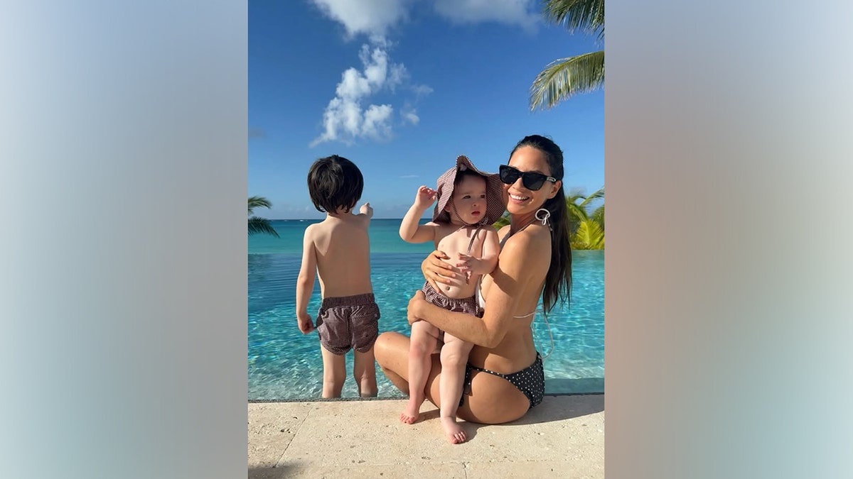 Olivia Munn poses with her children while on vacation by the pool as they play in the water.