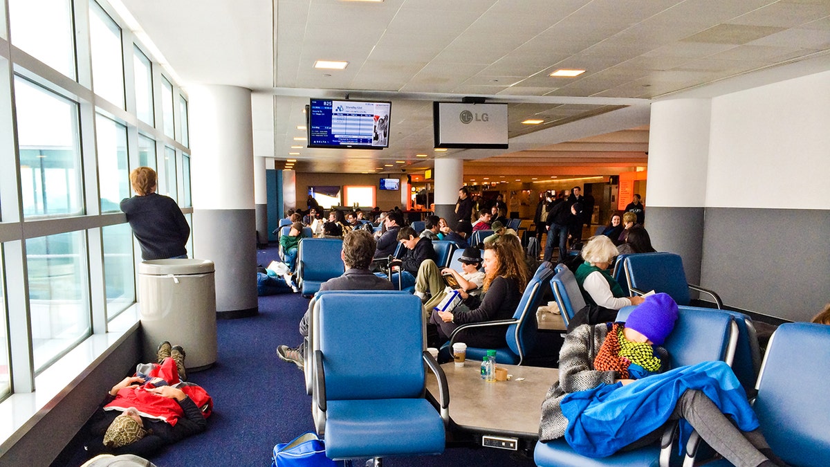 Busy airport gate area with travelers waiting, resting, and charging devices near large windows.