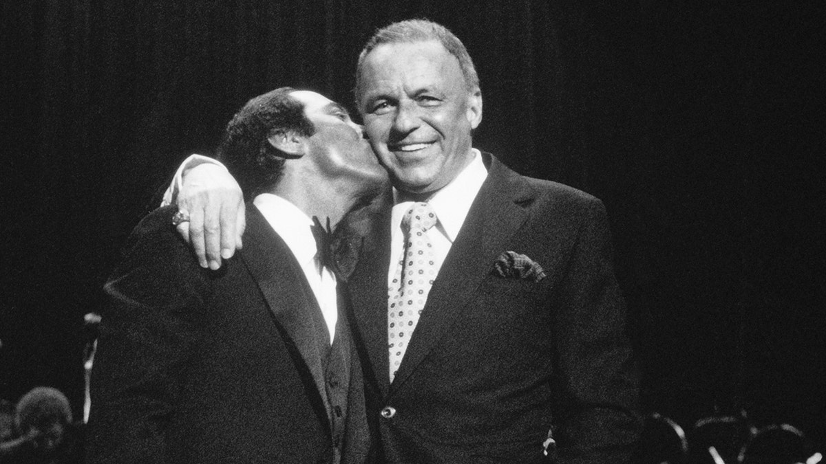 Paul Anka kissing a smiling Frank Sinatra on the cheek.