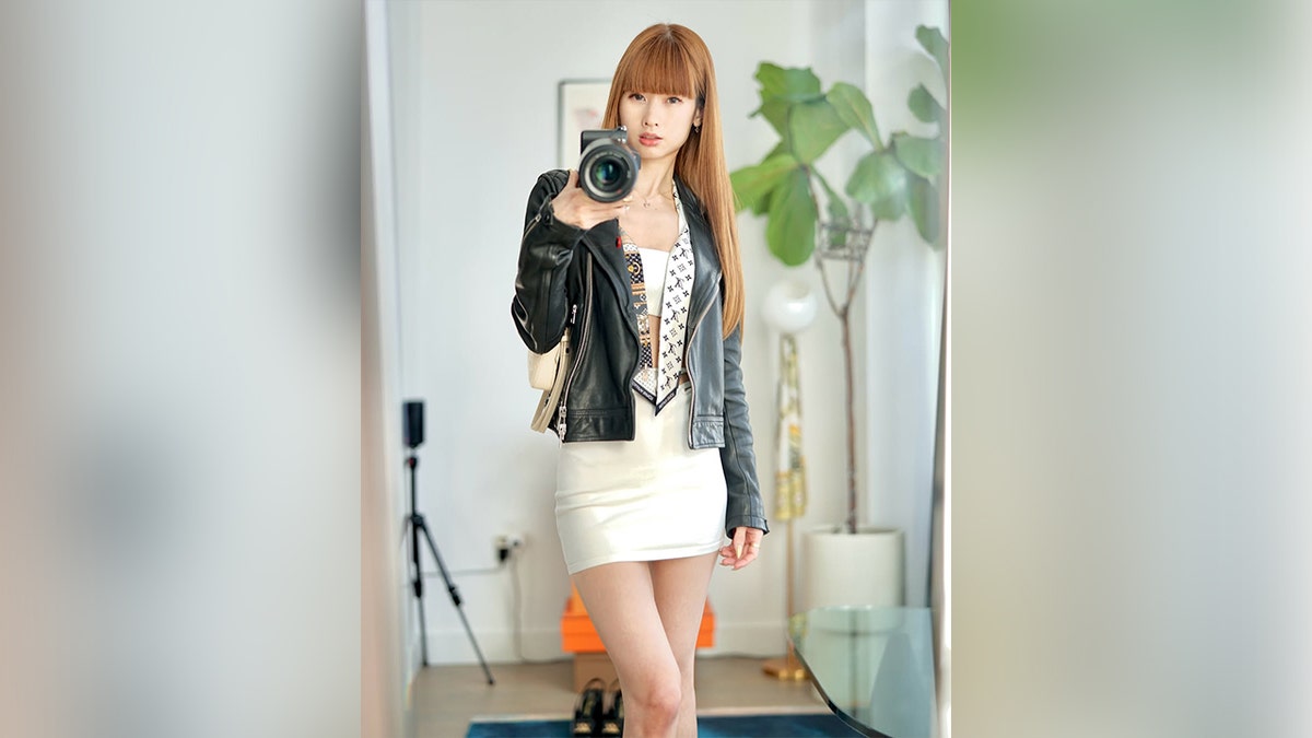 brunette woman with bangs in leather jacket and white skirt taking selfie in mirror of apartment