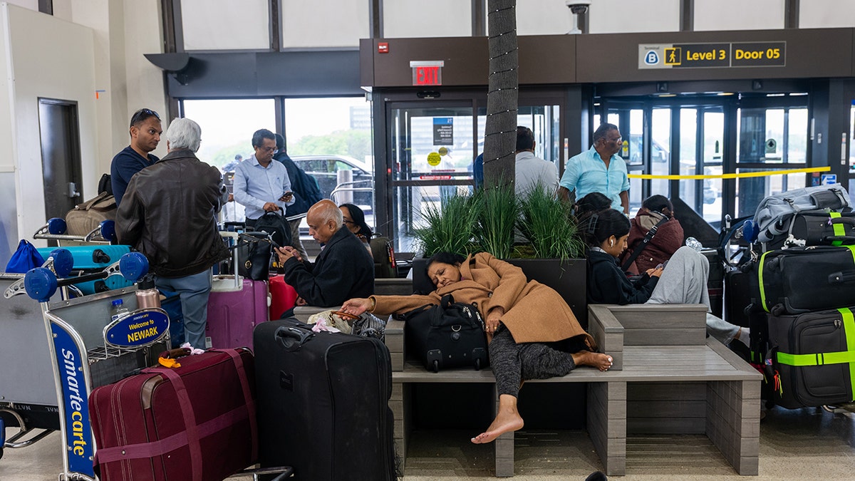 Um grupo de pessoas aguarda na saída do aeroporto, com uma mulher dormindo no banco.