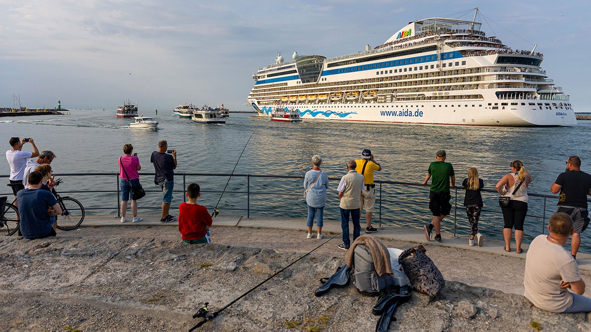 Large AIDA cruise ship departing a harbor as people watch and take photos from the waterfront promenade.