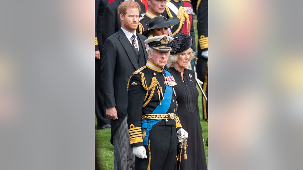 Prince Harry looking somber in a black suit as he stands behind King Charles and Queen Camilla.
