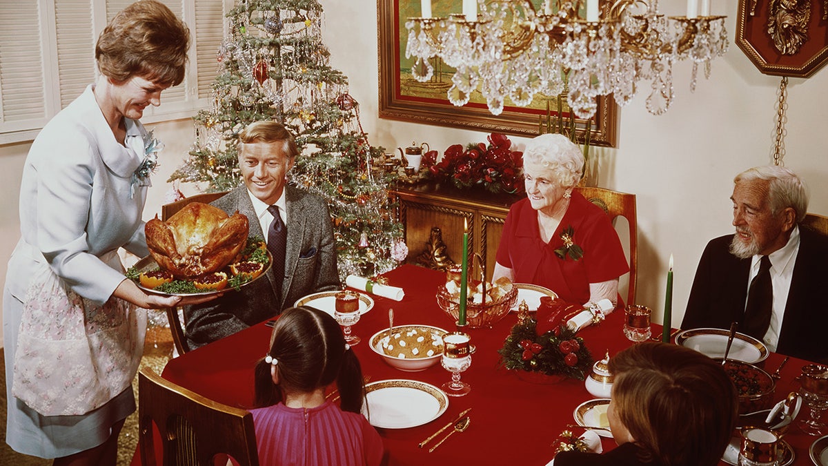 Family enjoying Christmas dinner in 196, watching as woman brings turkey to table to kids, husband and grandparents.