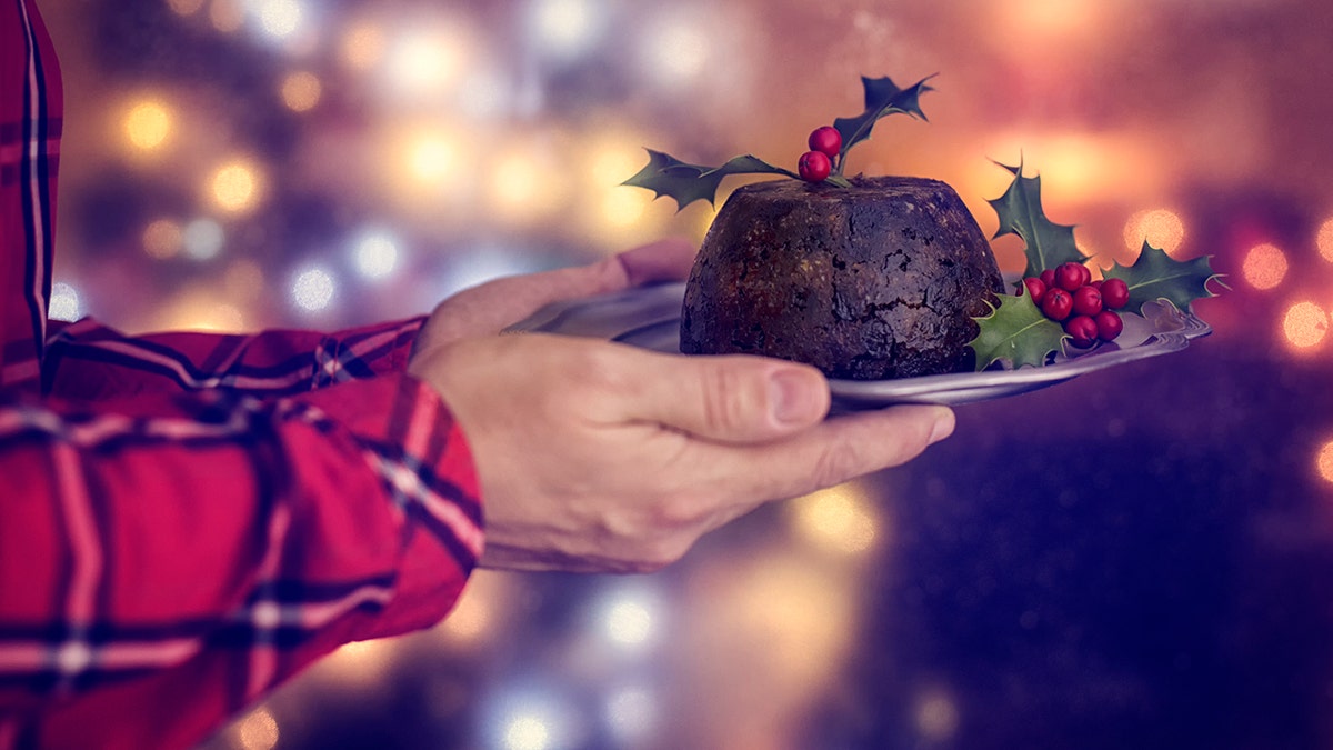 Man's hands holding plate of festive Christmas plum pudding with lights and decorations seen behind him.