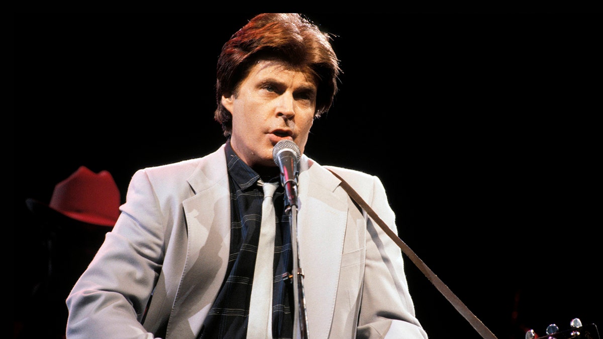 Ricky Nelson wearing a grey suit and playing guitar on stage.