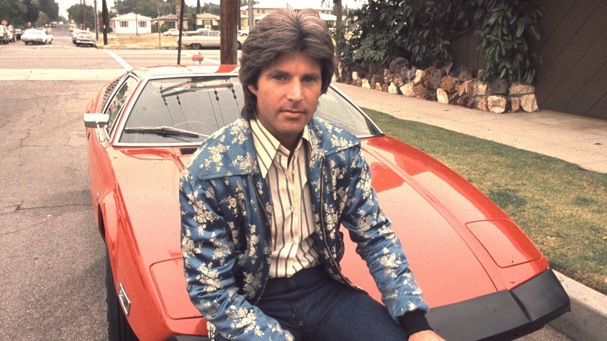 Ricky Nelson wearing a multiprinted jacket sitting in front a red car.