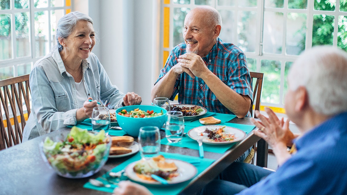 Senior woman and two senior men chatting, looking happy while eating healthy foods around table.