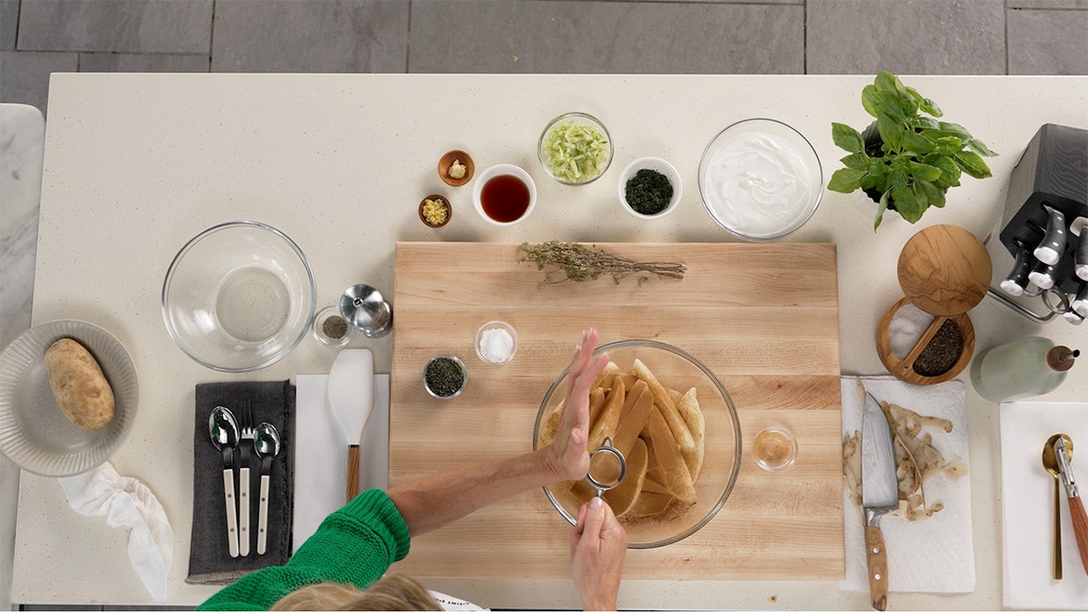 A bird's eye view of a chef pouring cinnamon on sliced potatoes in a glass bowl.