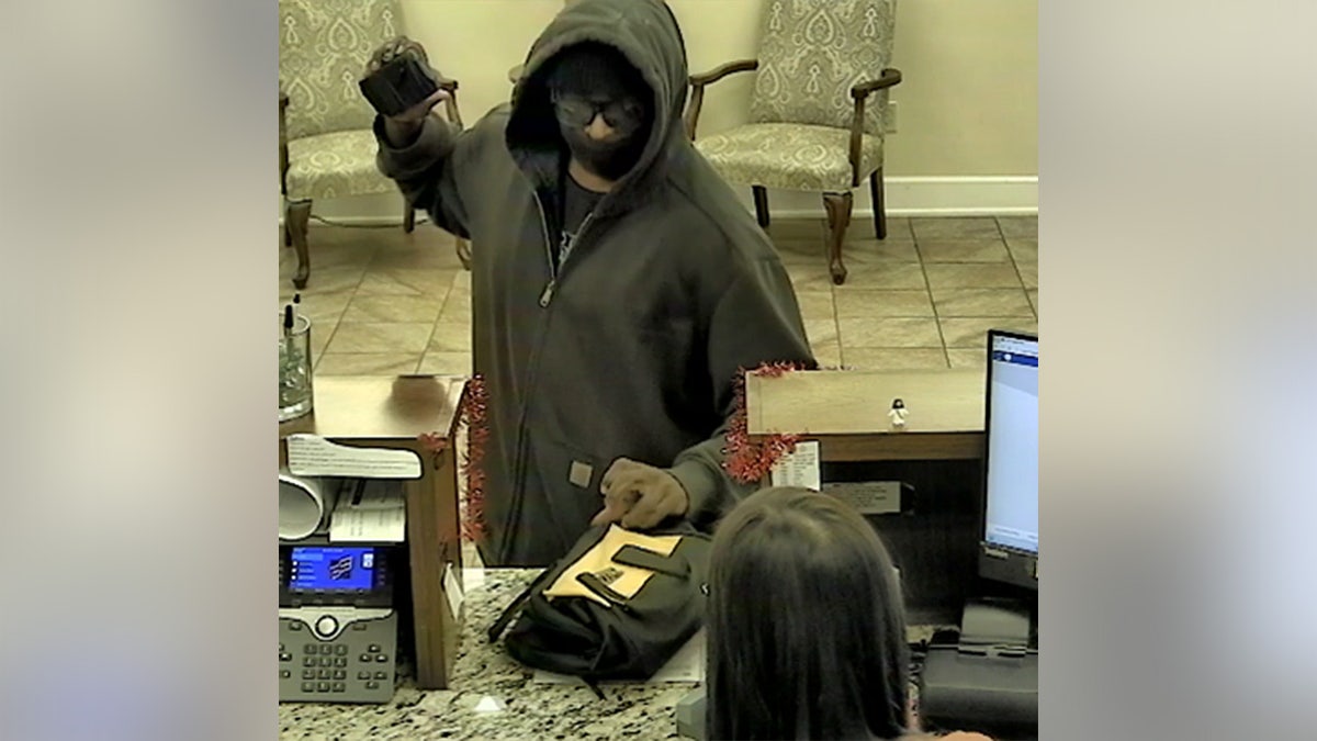Bank surveillance image shows a masked suspect wearing a hooded sweatshirt at a teller counter, holding an object in one hand while a bag rests on the counter between the individual and a bank employee.