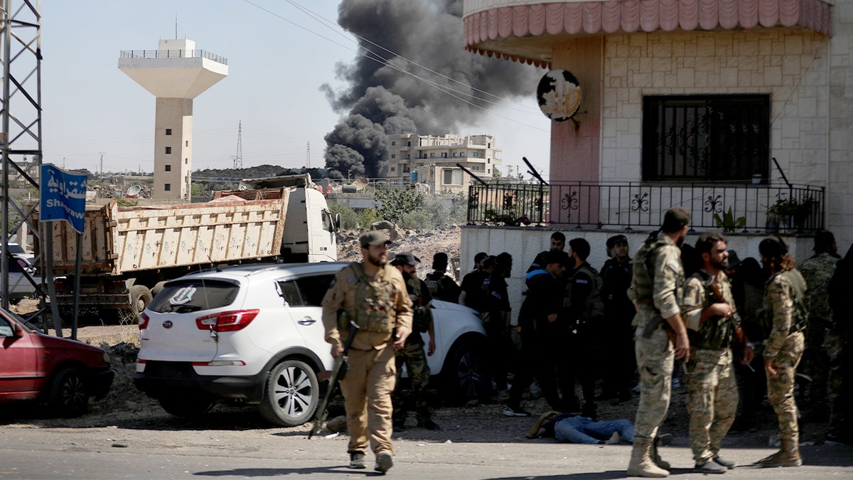Soldiers gather in a rural area as smoke rises in the distance from recent unrest.
