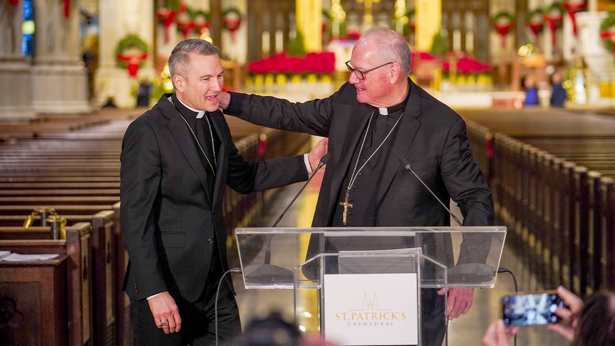 Two Catholic leaders shake hands during a formal gathering with media present.