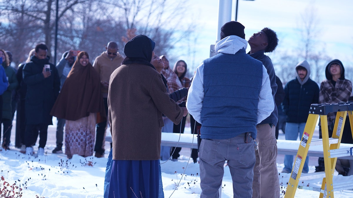 Crowd stands in snow looking up as flag is hoisted
