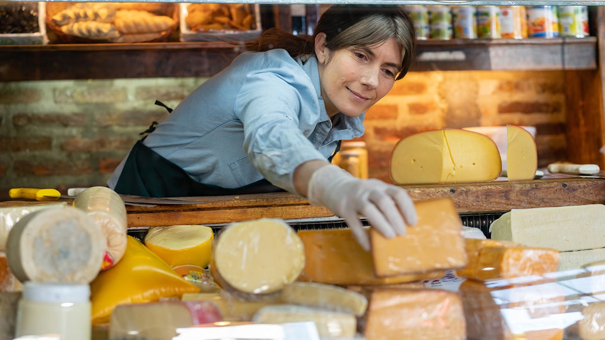 Female owner of a delicatessen arranging big chunks of cheese in a display seen from other side of the glass.