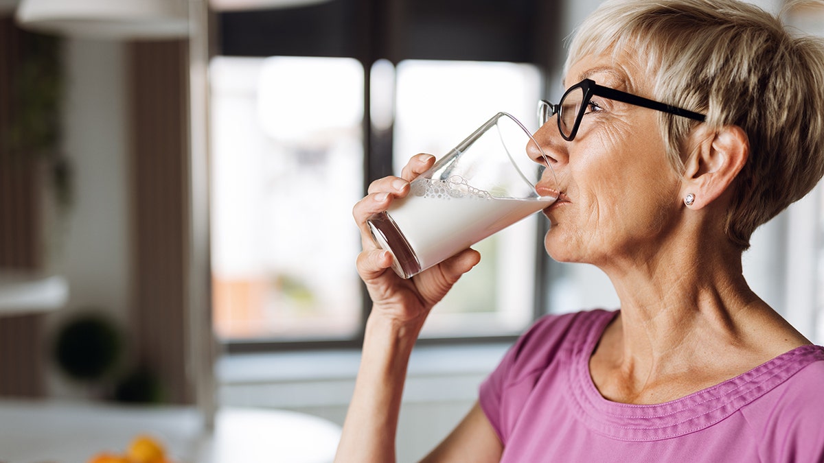 Older woman with blonde hair and black eye glasses is drinking milk.