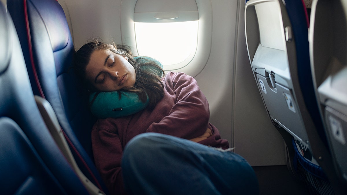 Airplane passenger sleeping in economy class window seat using a travel neck pillow.